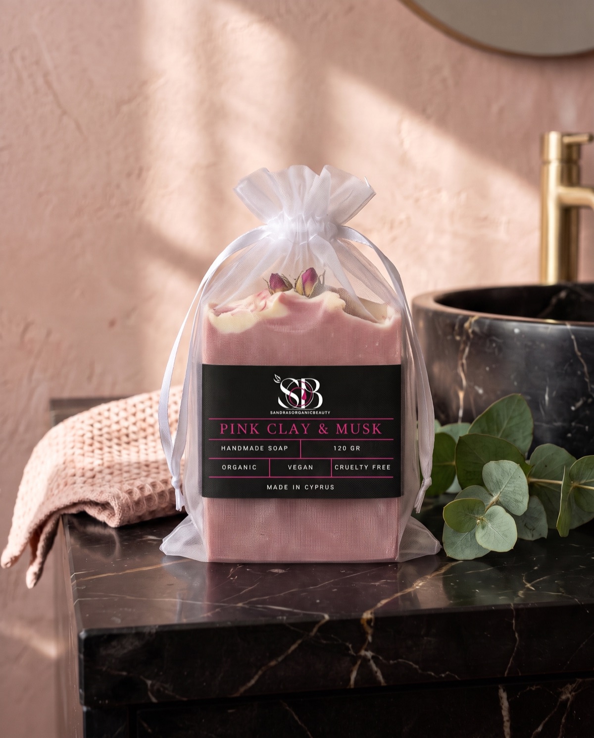A bar of pink clay and musk soap in a white organza gift bag beside a black marble basin
