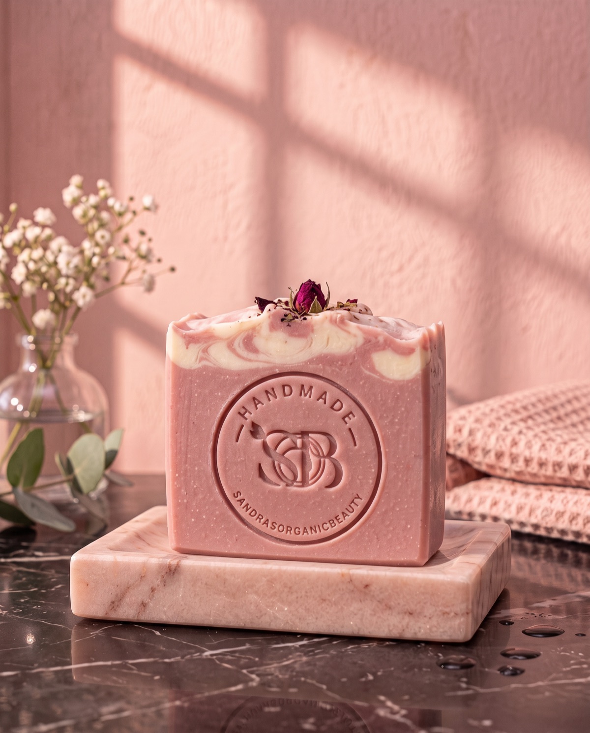 A bar of pink clay soap with a swirled top and a dried rosebud, stamped Handmade SOB, on a pink marble soap dish