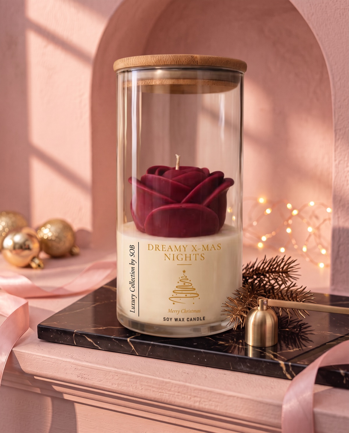 A deep red sculpted rose candle in a tall glass jar with a bamboo lid, labelled Dreamy X-Mas Nights, on a black marble board with gold baubles and fairy lights